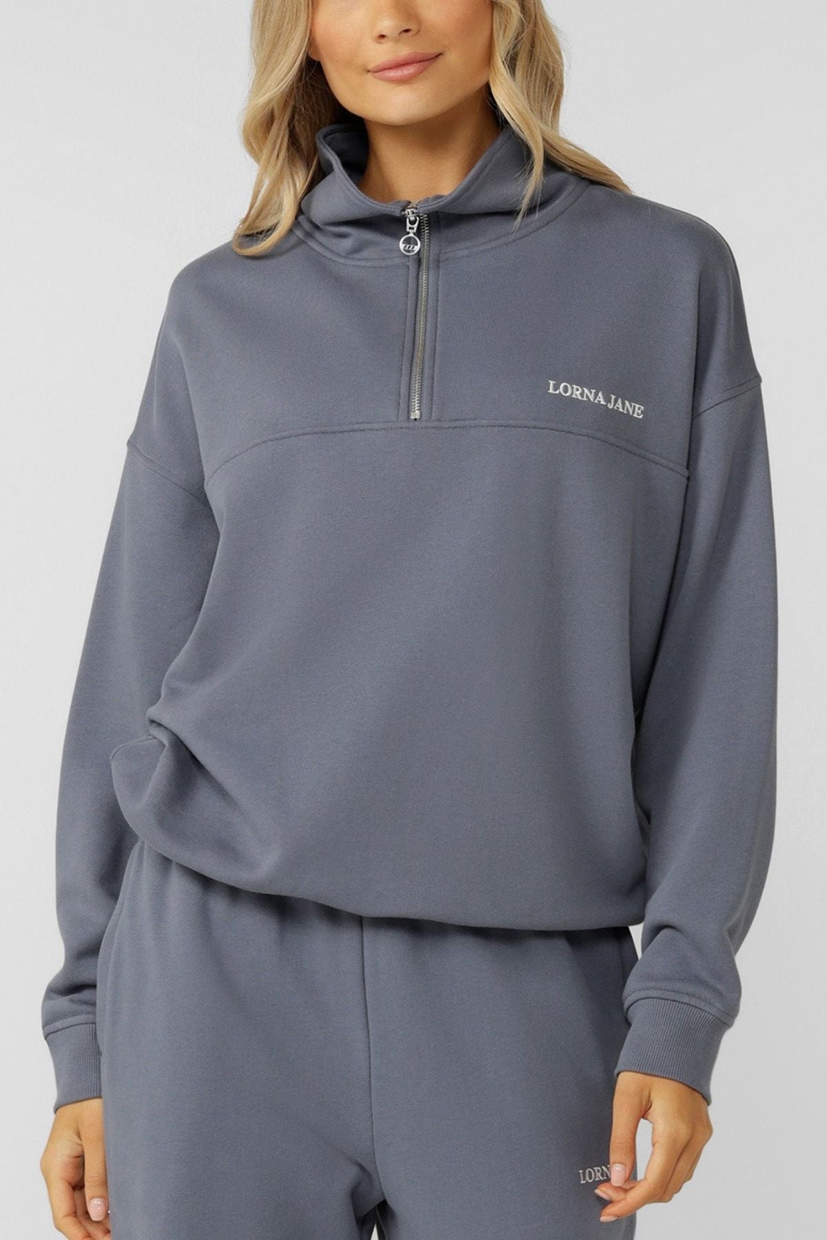 A woman wears the Lorna Jane Streamline Quarter Zip Sweat and matching sweatpants, both loose-fitting and gray with "LORNA JANE" branding. Made from soft cotton terry, she stands with one hand in her pocket against a plain background.