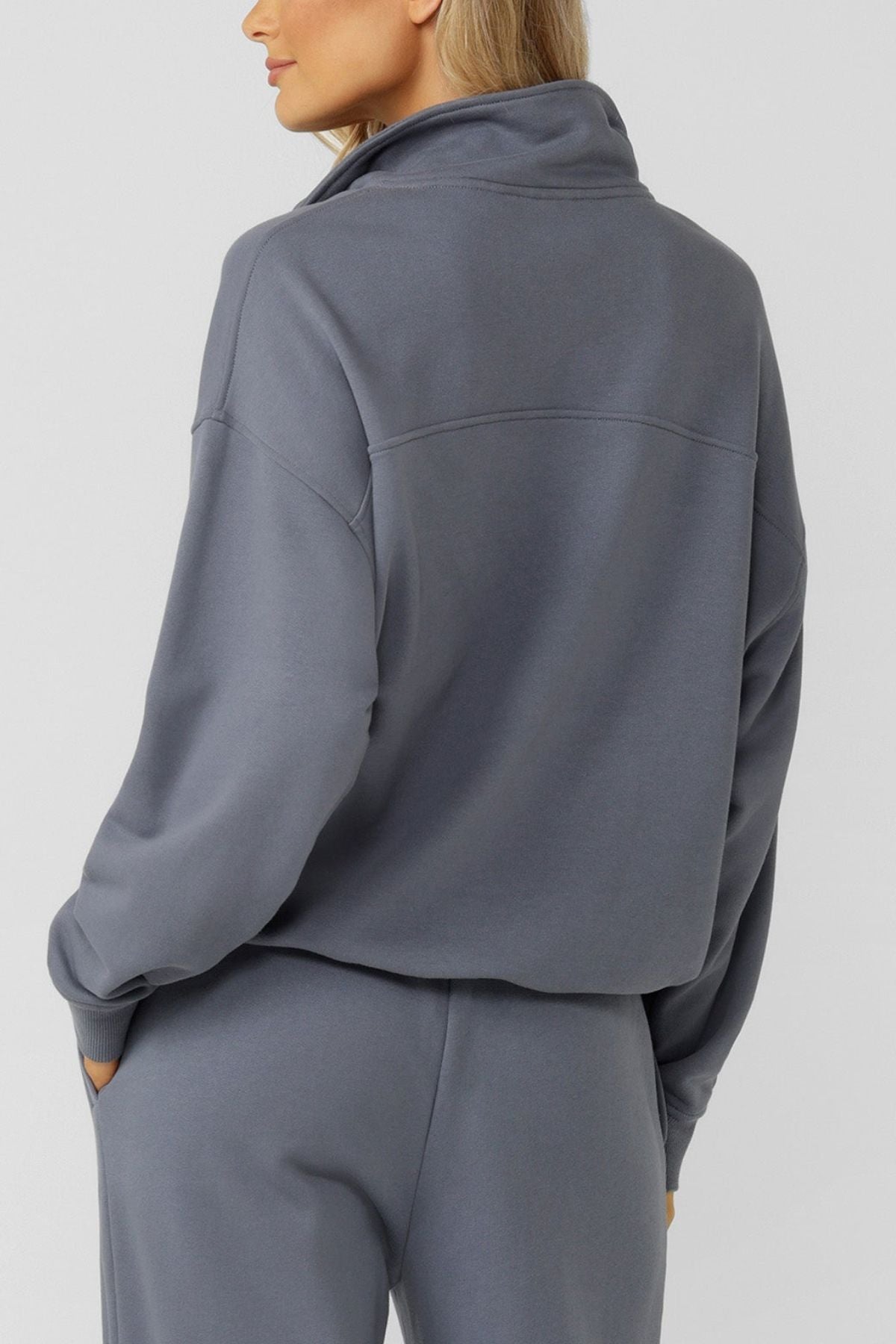 Someone wearing the Lorna Jane Streamline Quarter Zip Sweat in gray with matching loose-fit pants, made from soft cotton terry, stands hands-in-pockets, facing away from the camera against a plain light background.