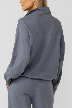 Someone wearing the Lorna Jane Streamline Quarter Zip Sweat in gray with matching loose-fit pants, made from soft cotton terry, stands hands-in-pockets, facing away from the camera against a plain light background.