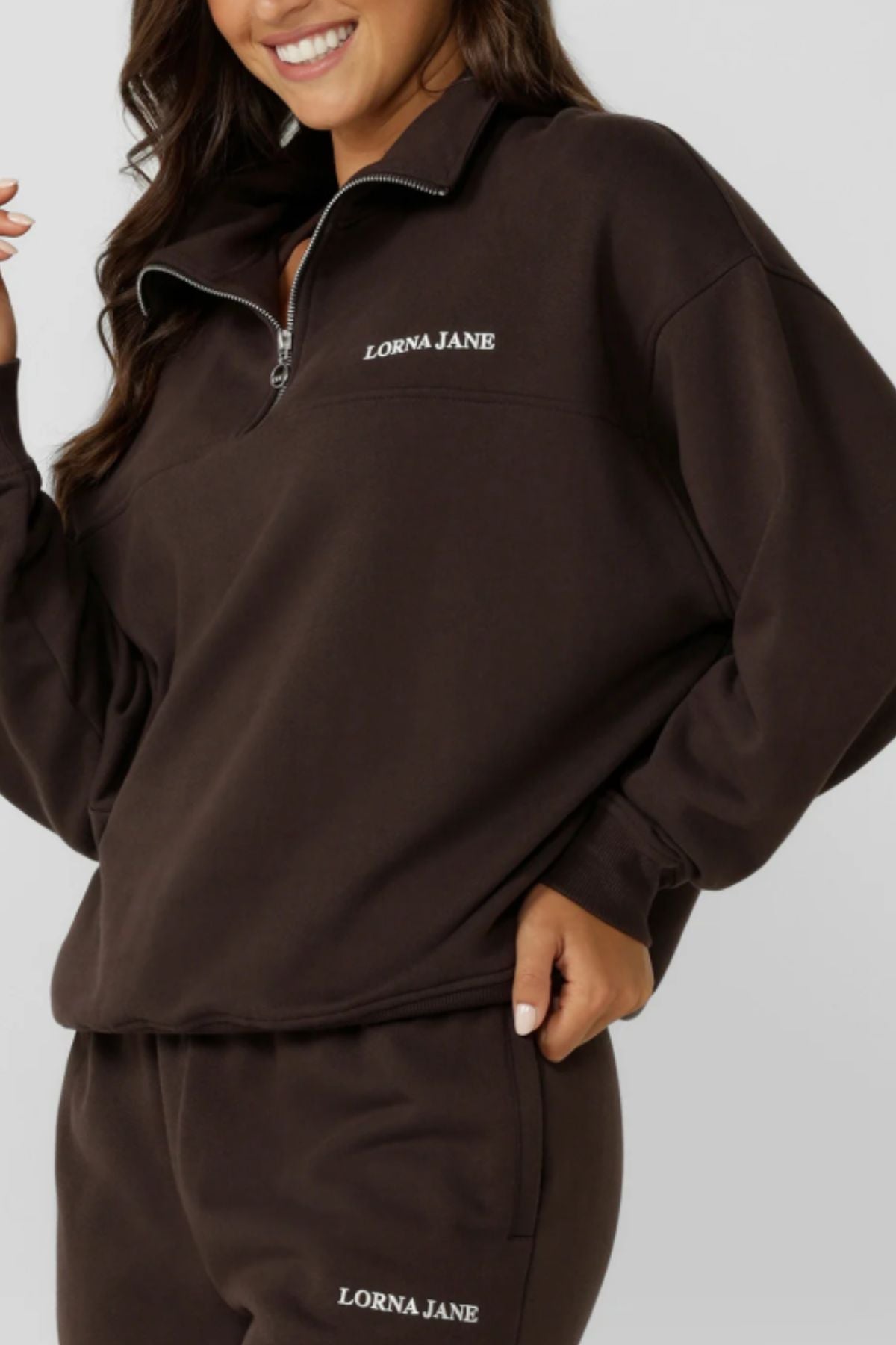 A woman smiles, wearing the Lorna Jane Streamline Quarter Zip Sweat in dark brown cotton terry with "LORNA JANE" in white, paired with matching sweatpants. Her hand rests on her hip, only her lower face and torso are visible.
