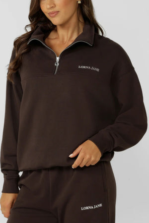 A woman wears the Lorna Jane Streamline Quarter Zip Sweat and matching pants in dark brown cotton terry, both labeled "LORNA JANE" in white. She poses against a plain light background—a perfect layering essential.