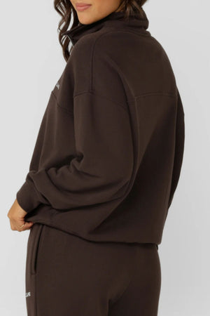 Wearing the Lorna Jane Streamline Quarter Zip Sweat in dark brown, a person stands sideways against a light gray background, gently pulling at the top for a cozy and relaxed look.