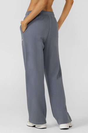A person stands with their back to the camera, wearing Lorna Jane Streamline Sweatpants in gray and white sneakers, one hand in their pocket. Their upper body is bare, highlighting part of their back and arm.