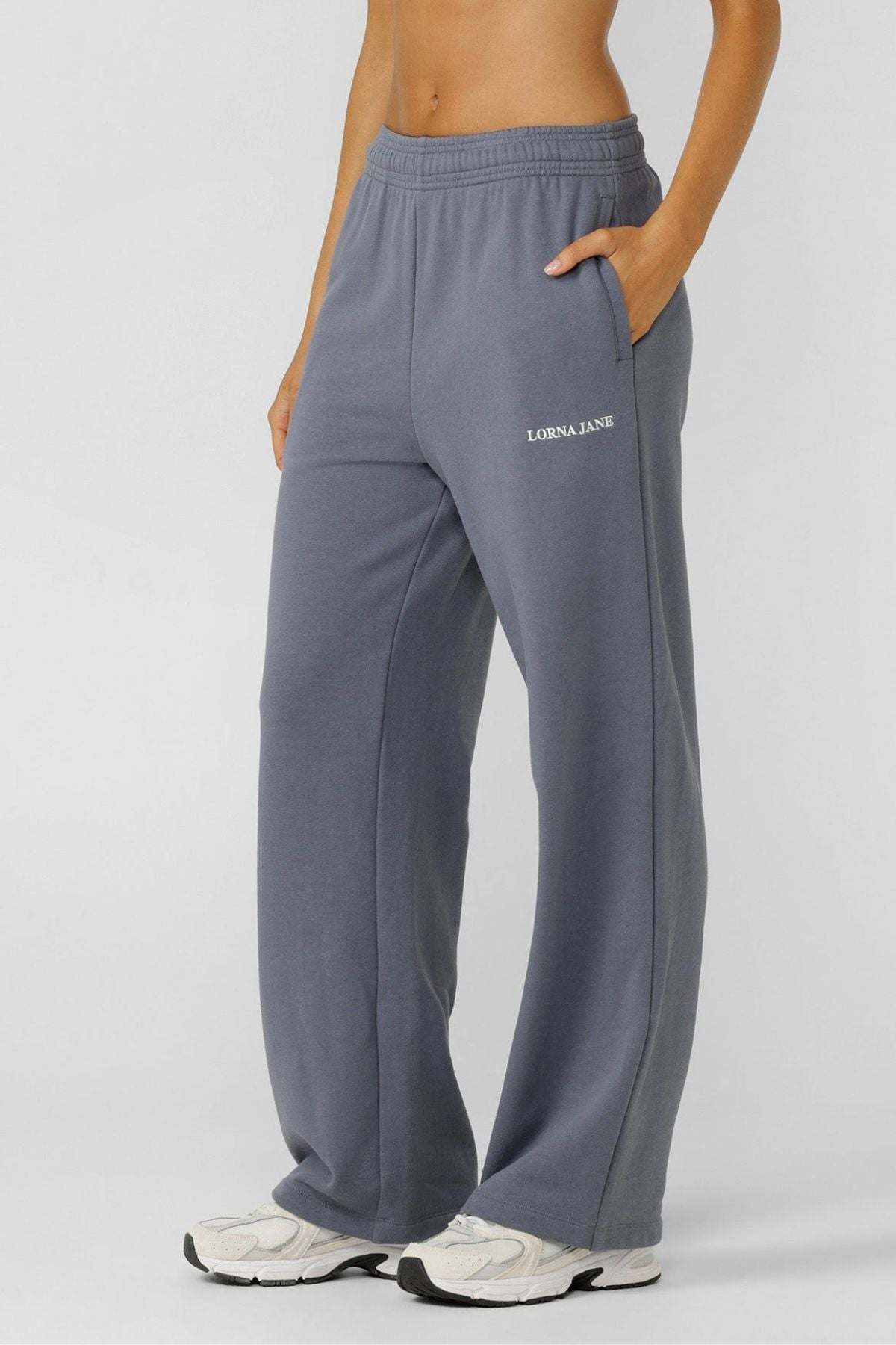 A person stands with one hand in the pocket of blue-gray, loose-fitting Lorna Jane Streamline Sweatpants and white sneakers against a plain background.