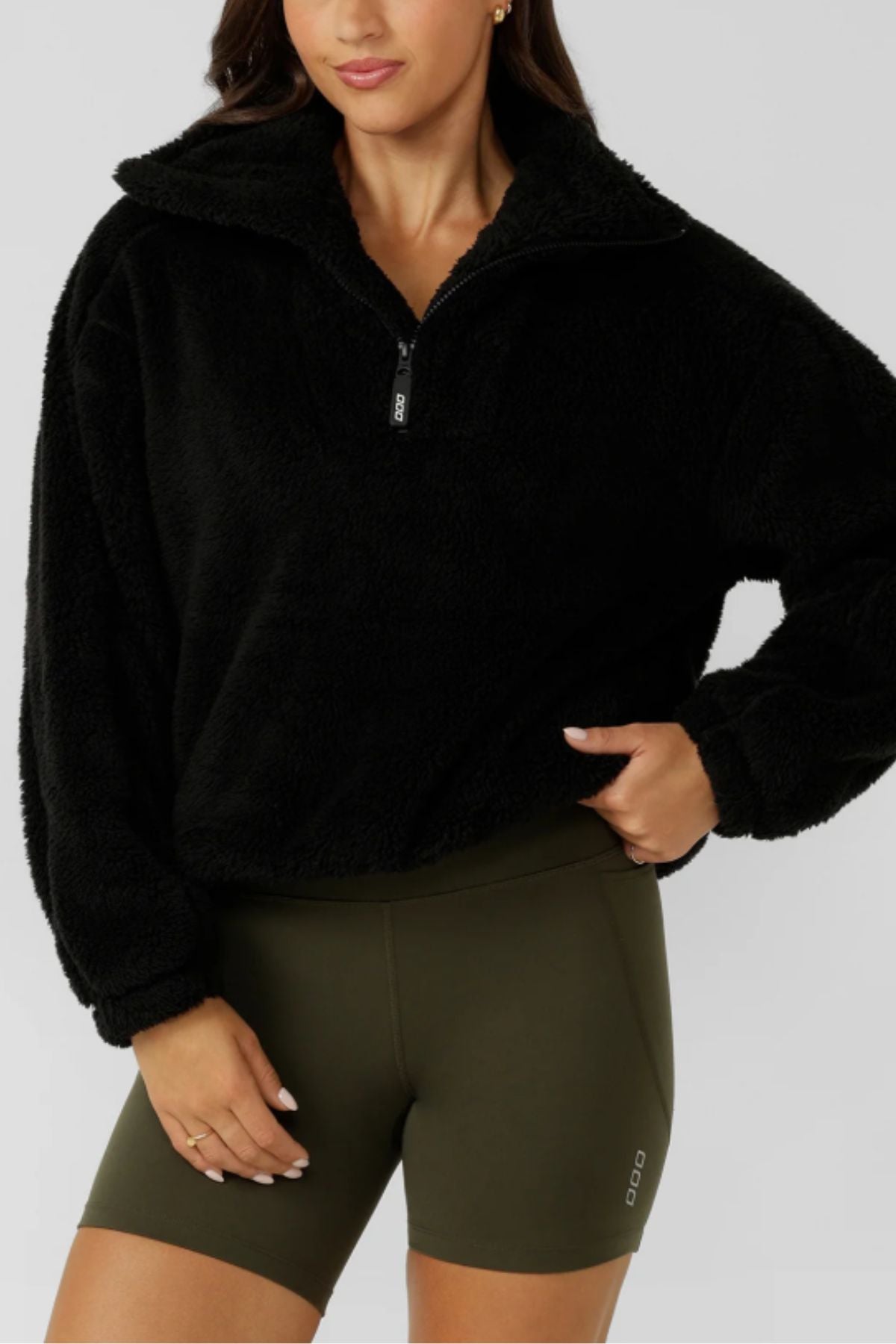 A woman wears the Lorna Jane Everyday Teddy Quarter Zip Sweat in black and olive green biker shorts, standing with one hand on her hip against a light gray background.