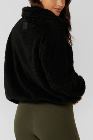 A woman models the Lorna Jane Everyday Teddy Quarter Zip Sweat, a black fuzzy oversized women's sweatshirt, paired with high-waisted olive green leggings, shown from the back and slightly turned to the side against a plain background.