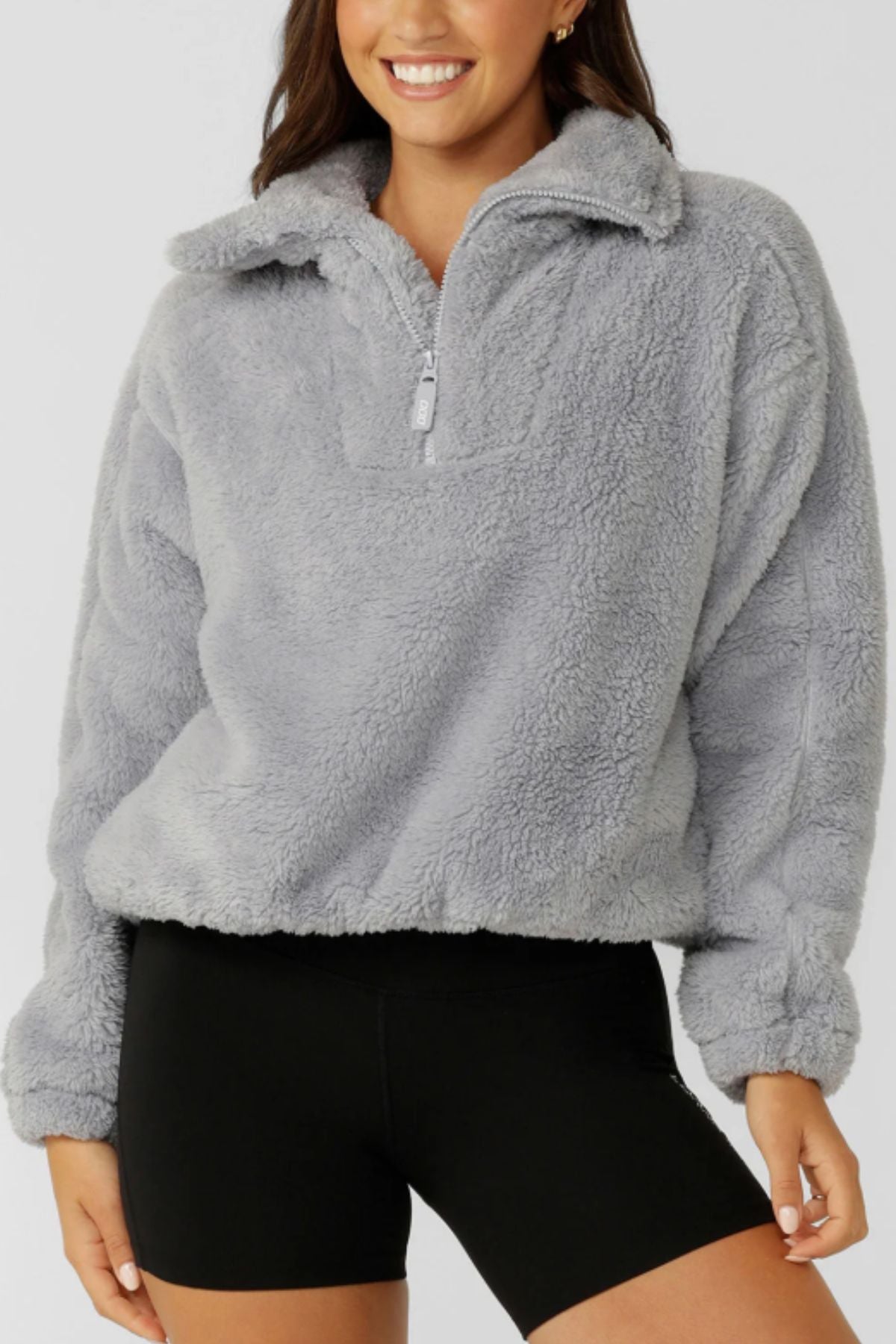 A person in a light gray Lorna Jane Everyday Teddy Quarter Zip Sweat and black fitted shorts smiles against a plain background.