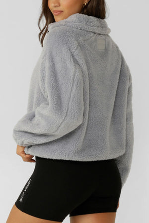 A person wearing the Lorna Jane Everyday Teddy Quarter Zip Sweat in light gray and black fitted shorts with "Lorna Jane" on the leg, posing with one hand on their hip against a plain background.