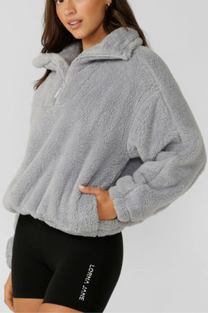 A woman wears the Lorna Jane Everyday Teddy Quarter Zip Sweat in light gray and black athletic shorts with white text on the left leg, resembling an oversized sweatshirt, against a plain light background.