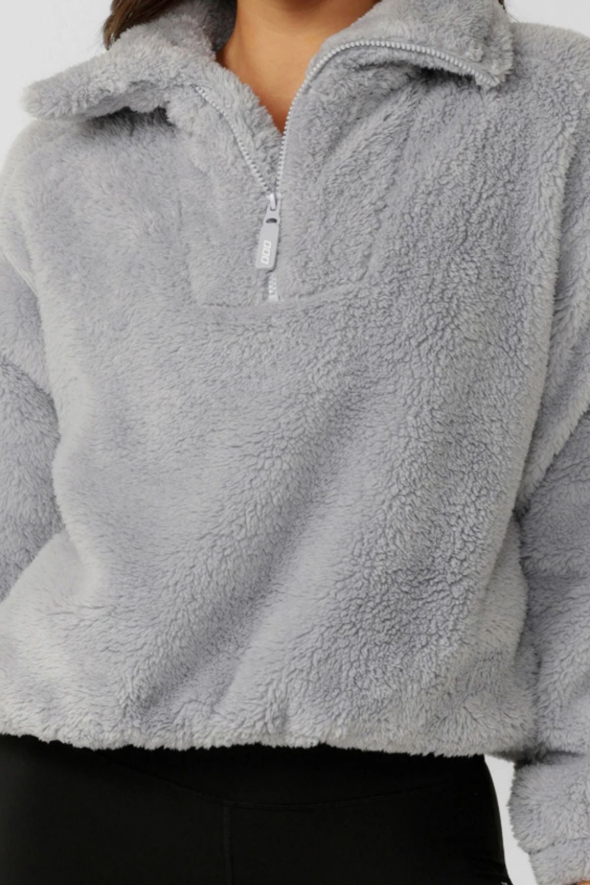 A person wearing the light gray Lorna Jane Everyday Teddy Quarter Zip Sweat with a high collar, paired with black pants. Only the torso and part of the face are visible.