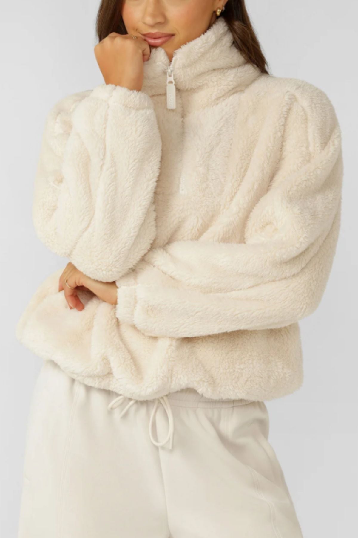 A person wearing a Lorna Jane Everyday Teddy Quarter Zip Sweat, a cosy oversized women's sweatshirt with a high collar, and matching cream drawstring pants stands with one hand touching their face.