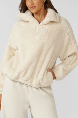 A woman models the comfy, cream Lorna Jane Everyday Teddy Quarter Zip Sweat with long sleeves and an oversized fit, paired with light pants. She stands against a plain background, one hand on her hip.