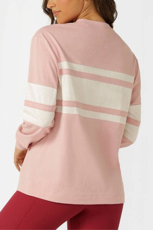 A woman wears the Lorna Jane Trackside Long Sleeve T-Shirt, featuring white stripes across the back and upper arms, paired with red leggings. She is turned slightly away from the camera.