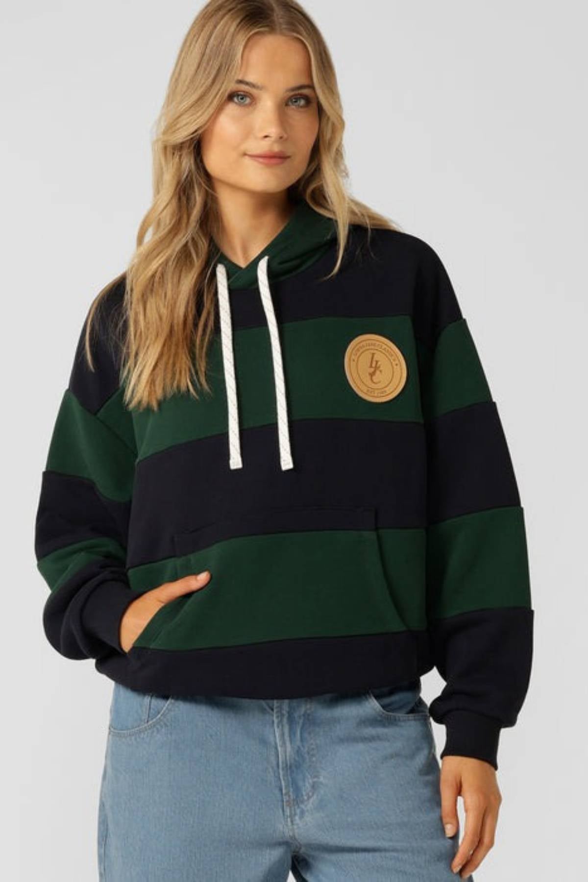 A woman with long blonde hair wears the Lorna Jane Heritage Rugby Polo Hoodie, featuring green and navy stripes with a beige chest patch, paired with light blue jeans. She stands against a plain background with one hand in the pocket, looking forward.