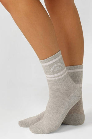 Someone wears Lorna Jane Lotus Crew Socks in light gray with white stripes and text on the upper cuff, standing on a white surface.