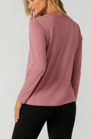 A blonde woman stands with her back to the camera, head turned slightly, wearing the Lorna Jane Lotus Long Sleeve T-Shirt in mauve and black pants, highlighting the activewear’s breathable fabric.
