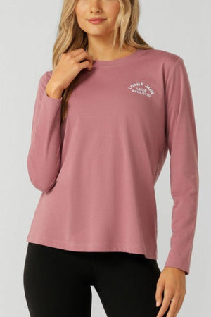 A woman in a Lorna Jane Lotus Long Sleeve T-Shirt (mauve) and black leggings stands before a plain light background, showcasing the breathable fabric and small white logo for a stylish, comfortable activewear look.