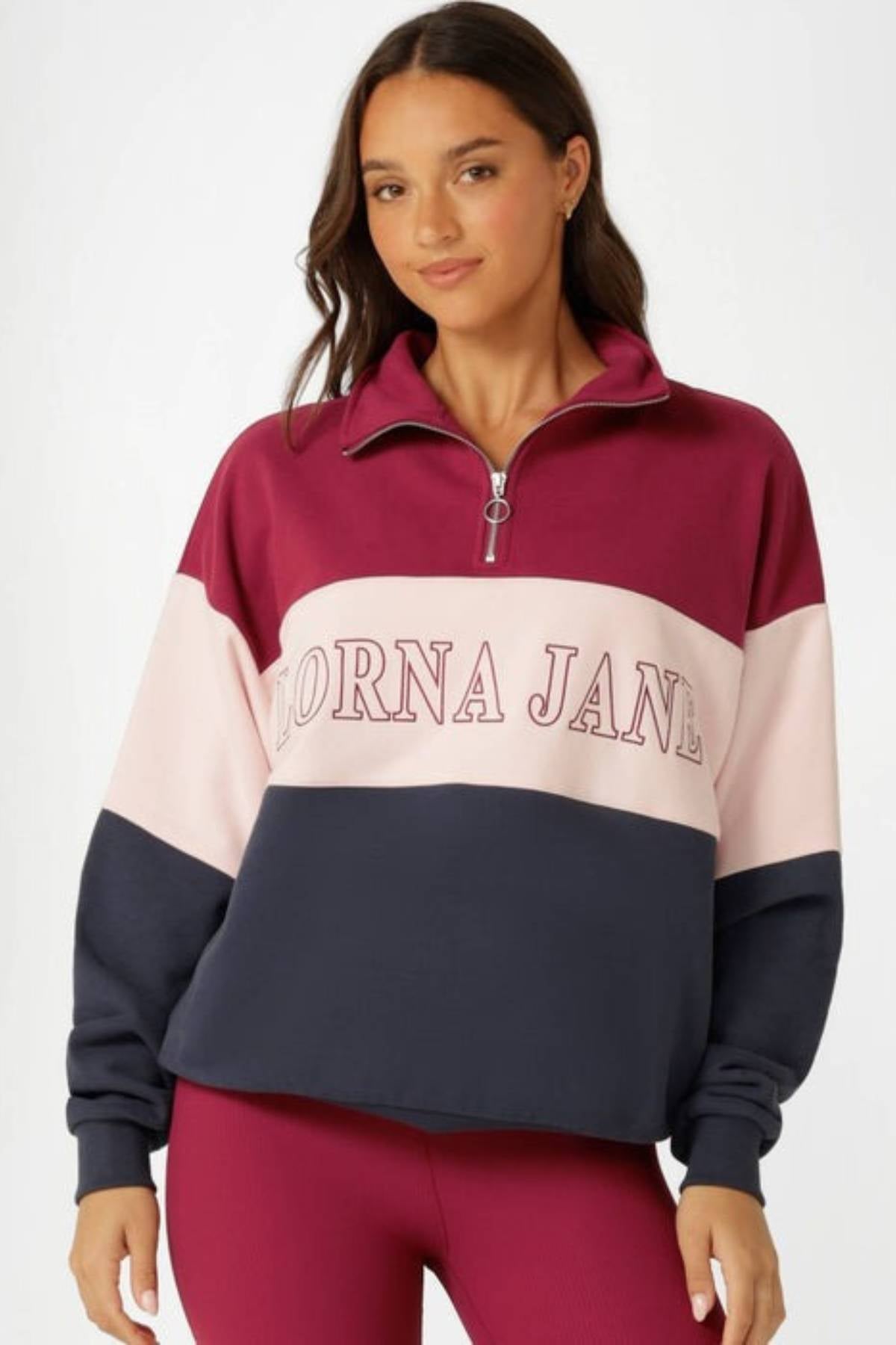 A woman wears the Lorna Jane Sidelines Panelled Quarter Zip Sweat—an oversized French terry cotton sweatshirt in burgundy, pink, and navy blue with "LORNA JANE" across the front—standing against a plain light background.