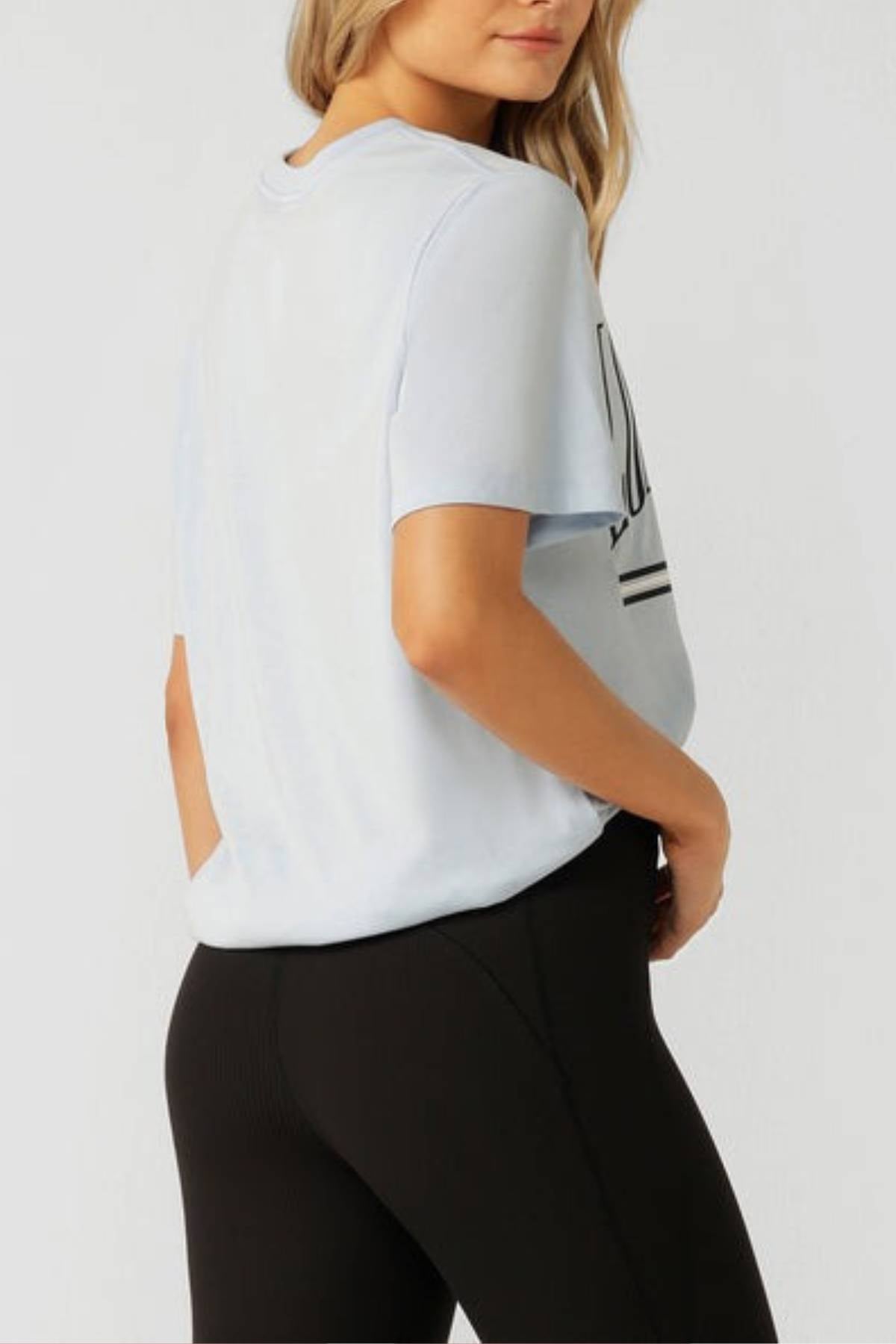 A woman stands sideways in a light blue Lorna Jane Topspin Relaxed T-Shirt and black leggings. Her left hand is behind her back, blonde hair loose and partly covering her face, set against a plain white background.