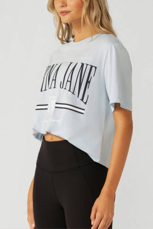 Woman wearing the Lorna Jane Topspin Relaxed T-Shirt in light blue with large black text and black high-waisted leggings, standing against a plain light background. Her face is partially out of frame.