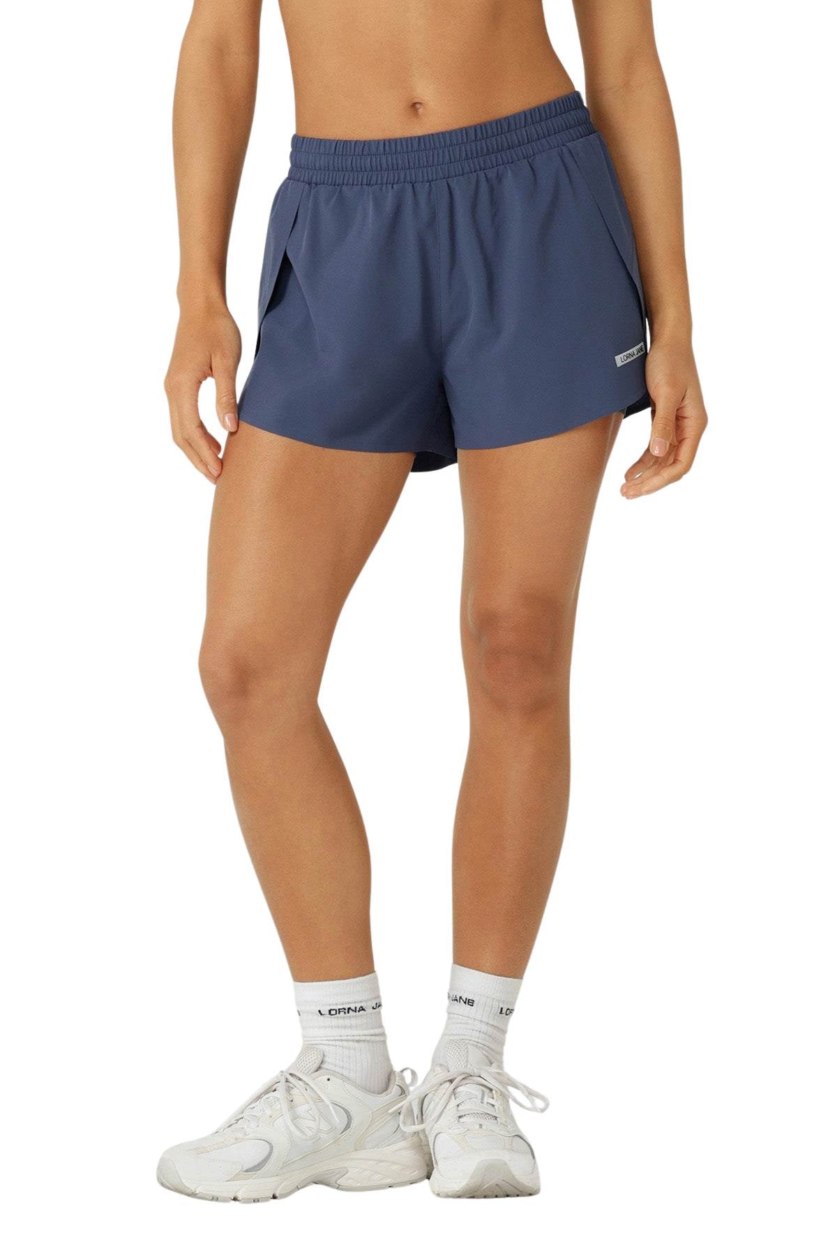 A person wears navy blue Lorna Jane Lightspeed Phone Pocket Run Shorts with anti-chafe lining, white socks, and sneakers, standing against a plain background. The photo is cropped from mid-torso to feet.