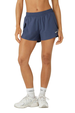 A person wears navy blue Lorna Jane Lightspeed Phone Pocket Run Shorts with anti-chafe lining, white socks, and sneakers, standing against a plain background. The photo is cropped from mid-torso to feet.