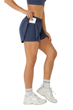 A person in a blue sports bra and Lorna Jane Lightspeed Phone Pocket Run Short stands sideways, placing a phone in the shorts’ pocket. They wear white socks and sneakers against a plain white background.