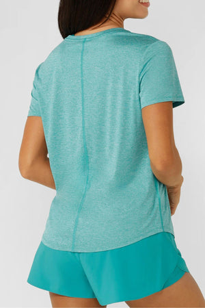 A woman, seen from the back, wears a light teal Lorna Jane Amy Dynamic Active T-Shirt and teal shorts. With one hand on her hip, she smiles slightly against a plain background. The workout top features breathable fabric for comfort.