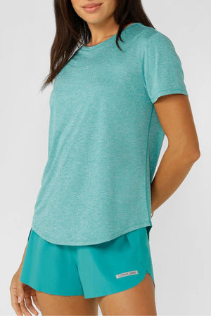 A woman’s torso and upper legs are shown as she models the Lorna Jane Amy Dynamic Active T-Shirt in light teal, paired with matching shorts. The stylish, breathable fabric and small brand label stand out against a plain light background.