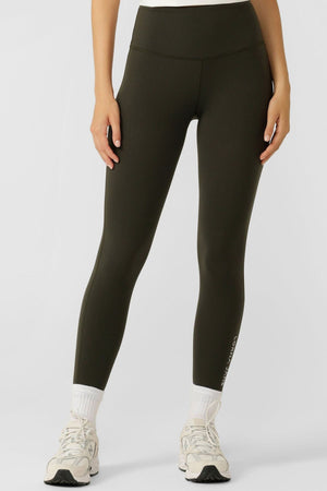 A person stands against a plain light background wearing Lorna Jane Lotus No Chafe Phone Pocket Ankle Biter Leggings in dark olive green, paired with white socks and white athletic shoes, highlighting trendy activewear. Upper body is cropped out.
