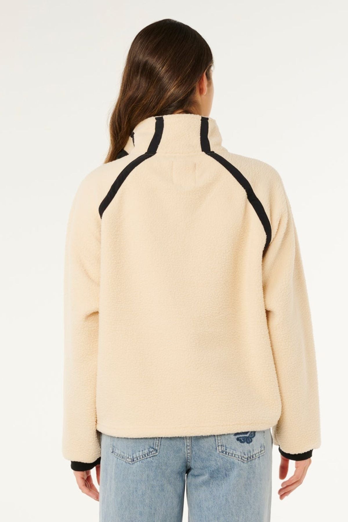 A person with long brown hair, viewed from behind, is wearing the Rip Curl Surf Club Heritage Polar jacket in cream with black trim, paired with light blue jeans. The background is plain white.