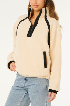 A person stands against a white background wearing the Rip Curl Surf Club Heritage Polar fleece pullover in light beige with black trim and zipper, paired with light blue jeans. Their left hand rests in the pocket, and their face is partially visible.