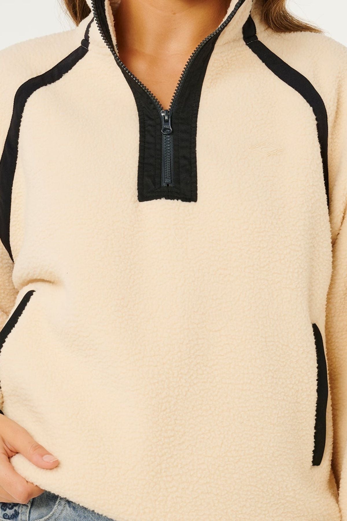 A person wearing the Rip Curl Surf Club Heritage Polar, a cream-colored fleece pullover with black trim, classic surf style design, zippered collar, and side pockets. Only the torso and part of one hand are visible.
