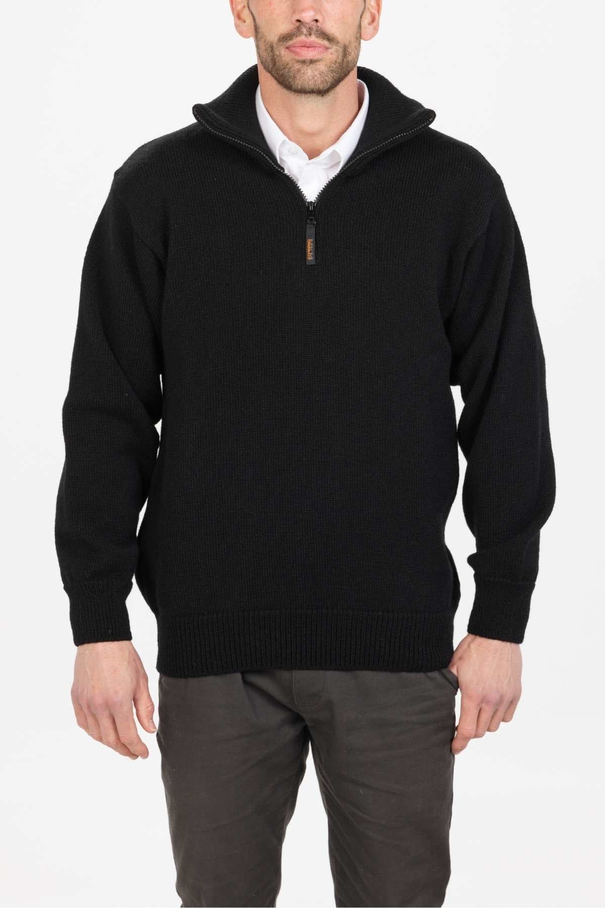 Dressed in the MKM North Wester Knitted Jumper, a black pullover made in New Zealand from 100% wool, paired with a crisp white collared shirt and gray pants, a man stands against a plain white backdrop, exuding style and quality craftsmanship.
