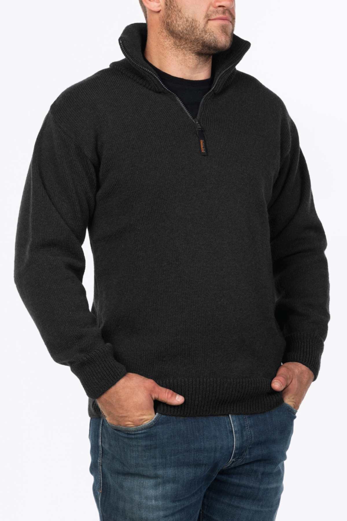 A man in a black MKM North Wester Knitted Jumper, made from 100% wool with a quarter-zip and high collar, stands with hands in his pockets wearing blue jeans. Made in New Zealand, he gazes to his right against a plain white background.