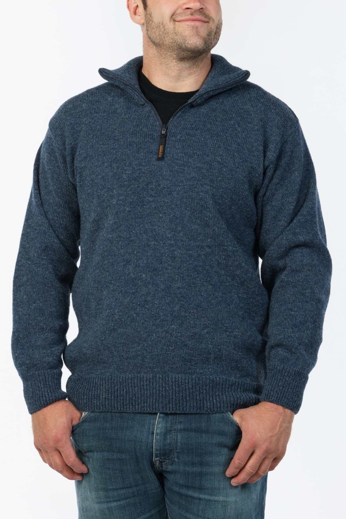 A man stands wearing the MKM North Wester Knitted Jumper and blue jeans, epitomizing casual wear. His hands are in his pockets, and he is smiling against a plain white background. The jumper, made in New Zealand, reflects both style and quality.