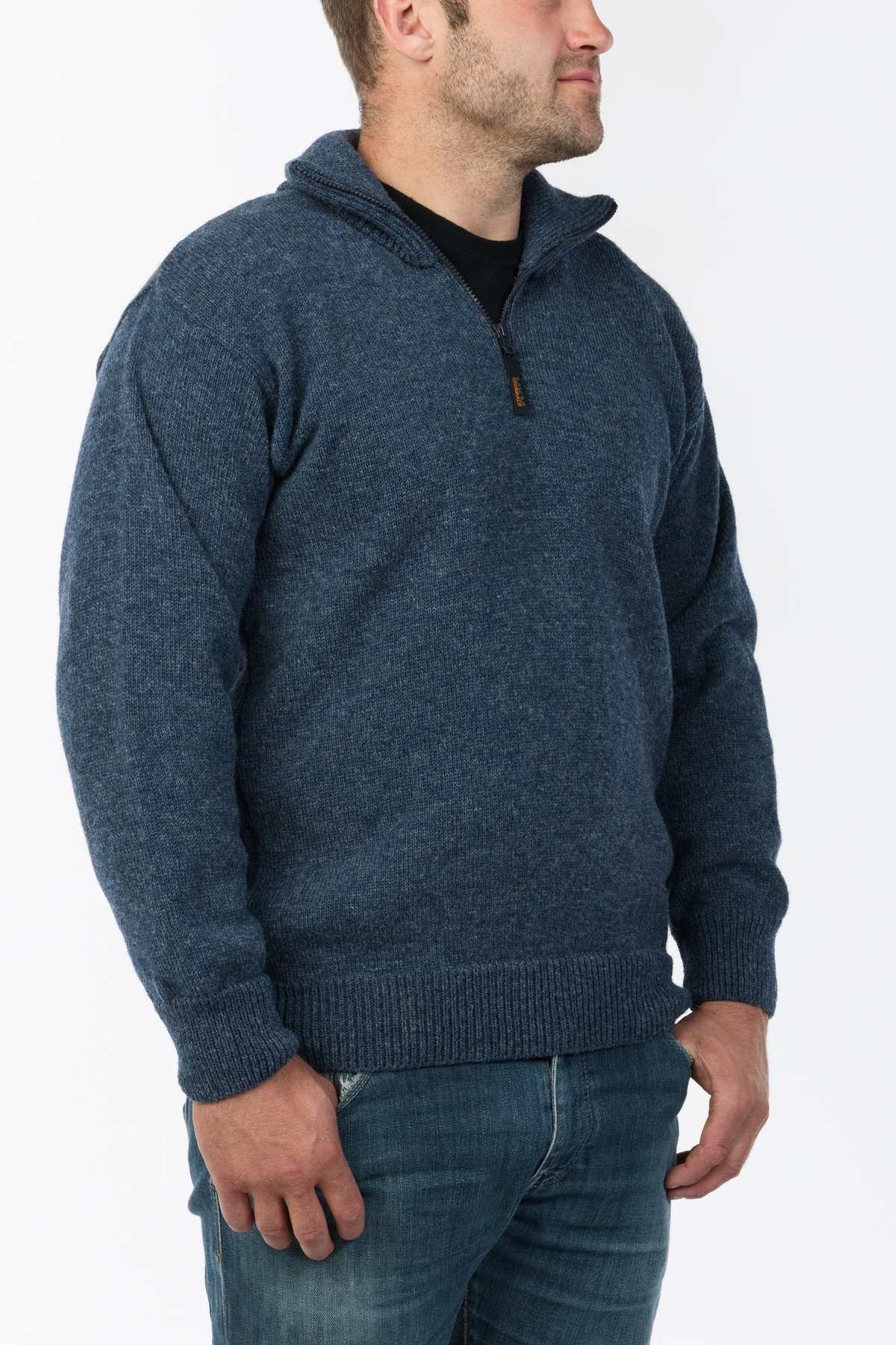 A man wearing the MKM North Wester Knitted Jumper in blue wool and blue jeans stands against a plain white background. He is facing slightly to the right with his hands relaxed by his sides. The jumper, made in New Zealand, features a ribbed collar, cuffs, and hem.
