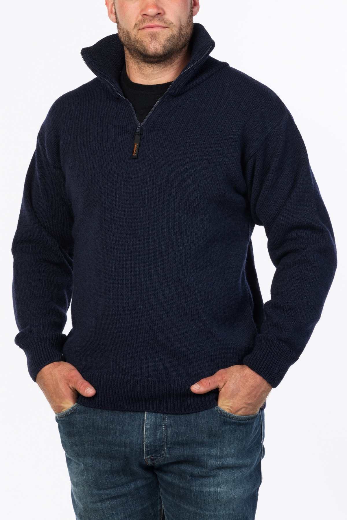 A person is wearing the MKM North Wester Knitted Jumper, a navy blue, half-zip sweater made in New Zealand from 100% wool. The high-collared sweater is layered over a black shirt, and their hands are in the pockets of their blue jeans. They are standing against a plain white background.