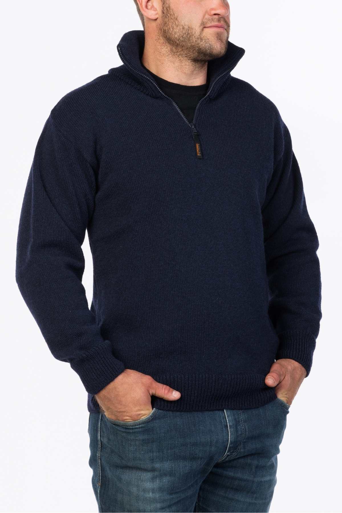A man wearing the MKM North Wester Knitted Jumper in navy blue, made from 100% wool, featuring a high collar and a half-zip design, along with blue jeans. He is standing with his hands in his pockets against a plain white background.