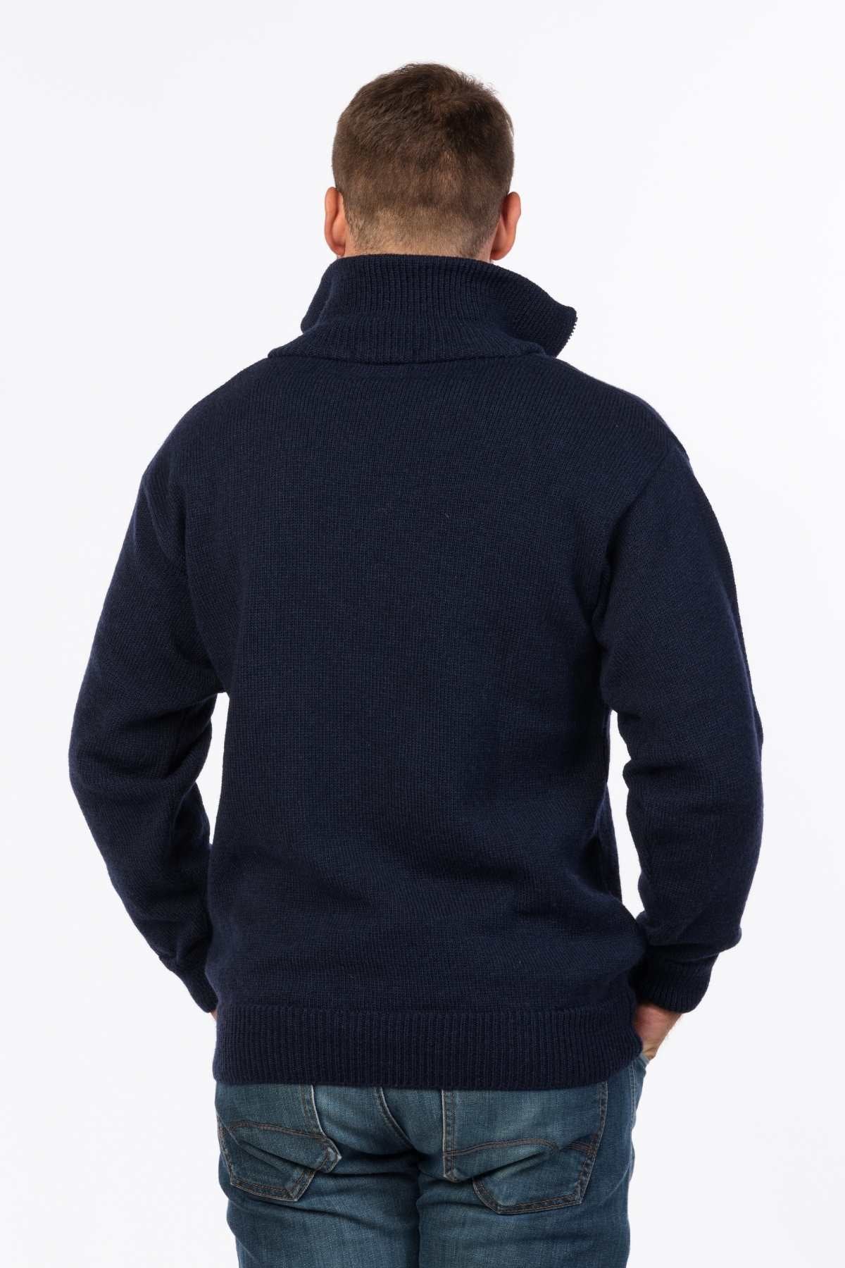 A person with short hair stands facing away from the camera, wearing a navy blue MKM North Wester Knitted Jumper made in New Zealand with the collar turned up and blue jeans. The person's right hand is in their pocket. The background is plain white.