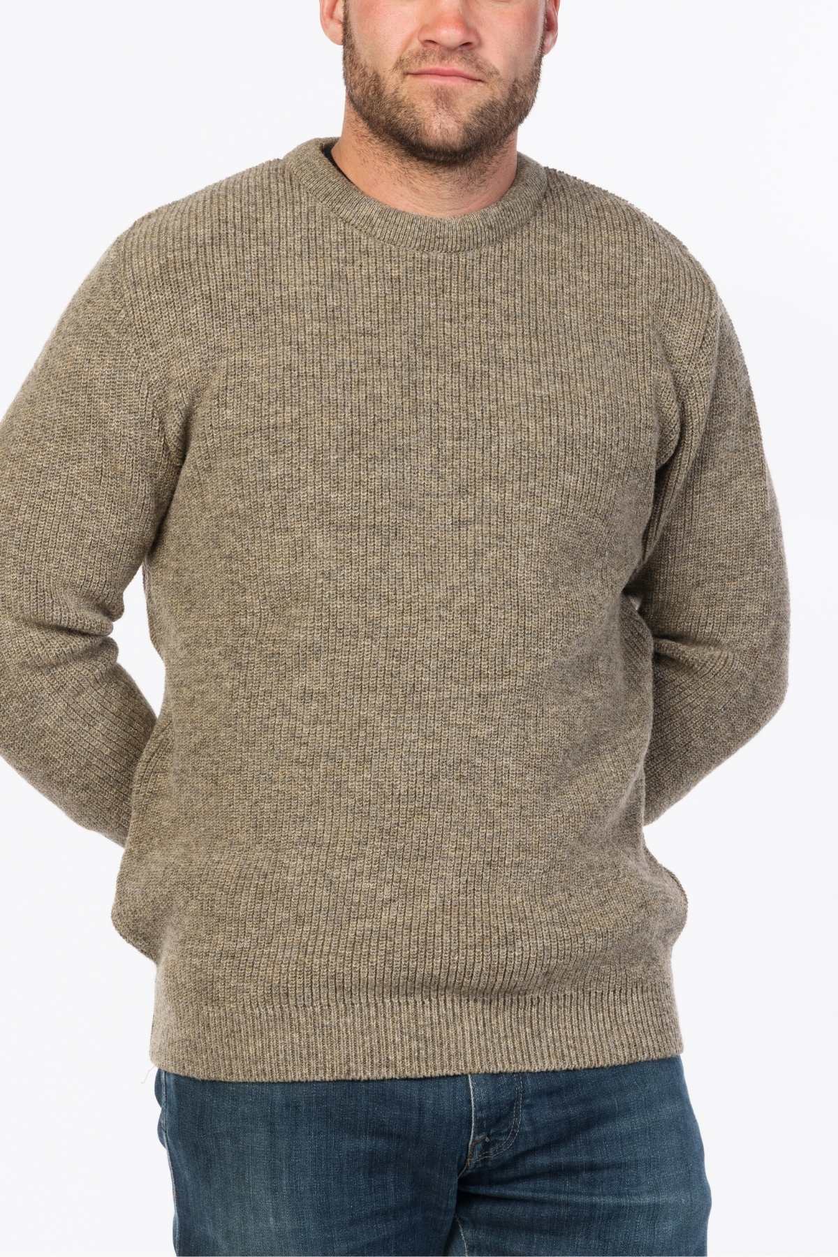 A person in the MKM Original Backyard Knit, a beige wool pullover, and blue jeans stands with their hands behind their back, set against a plain white background highlighting the simplicity of their casual look.
