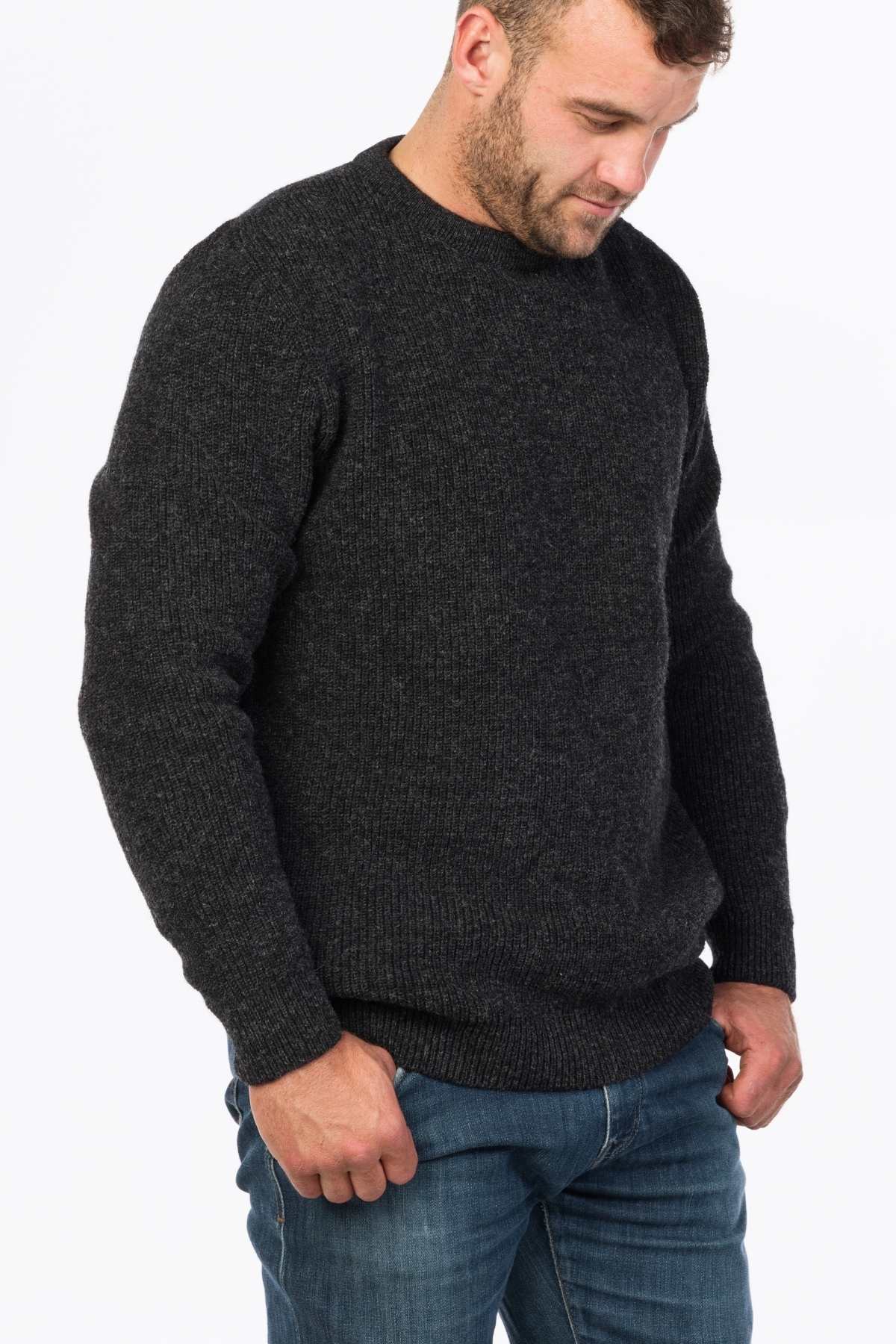 A man in a dark gray wool MKM Original Backyard Knit and blue jeans stands with hands in pockets, looking down. This casual look is both relaxed and stylish against the white background.