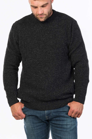 A man in an MKM Original Backyard Knit dark gray wool pullover, crafted in New Zealand, and blue jeans stands against a plain light background, looking down with hands in pockets.