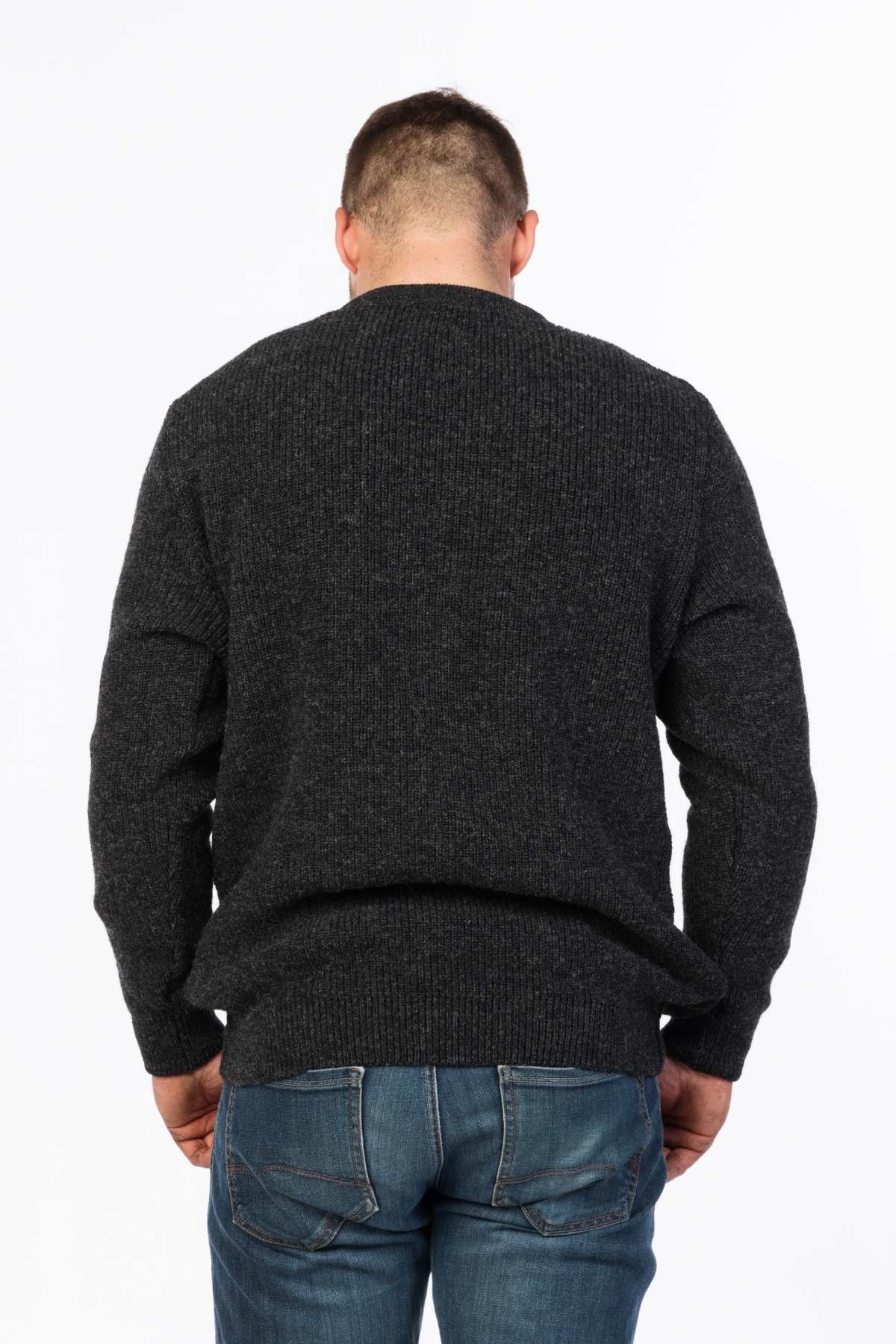 A man stands with his back to the camera, casually dressed in an MKM Original Backyard Knit dark gray wool pullover made in New Zealand and blue jeans, against a plain white background.