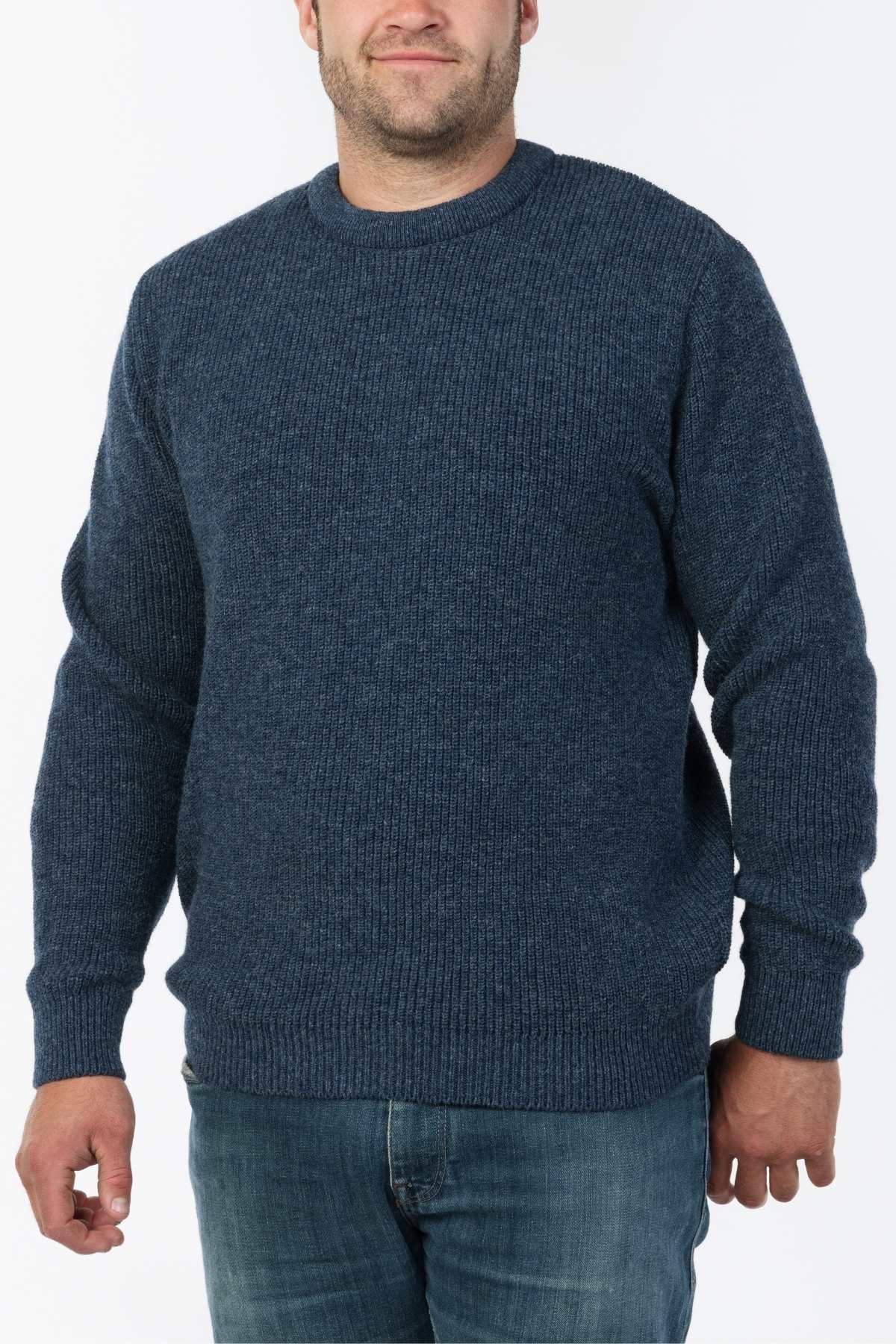 A person dons the MKM Original Backyard Knit, a blue knitted sweater with a ribbed texture and crew neckline, paired with blue jeans—a classic example of New Zealand's casual wear style against a plain white backdrop.