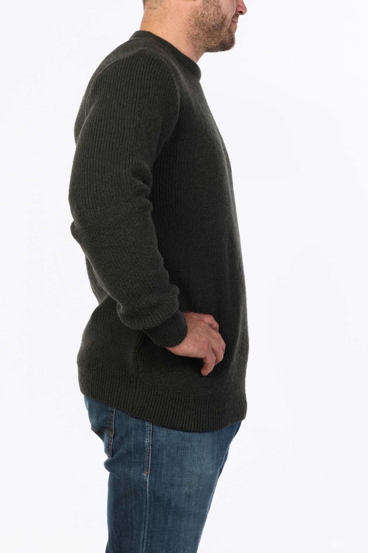 Side view of a person wearing the MKM Original Backyard Knit in dark green wool and blue jeans, epitomizing casual comfort. Crafted with quality in New Zealand, they stand with their right hand on their hip against a plain white background.