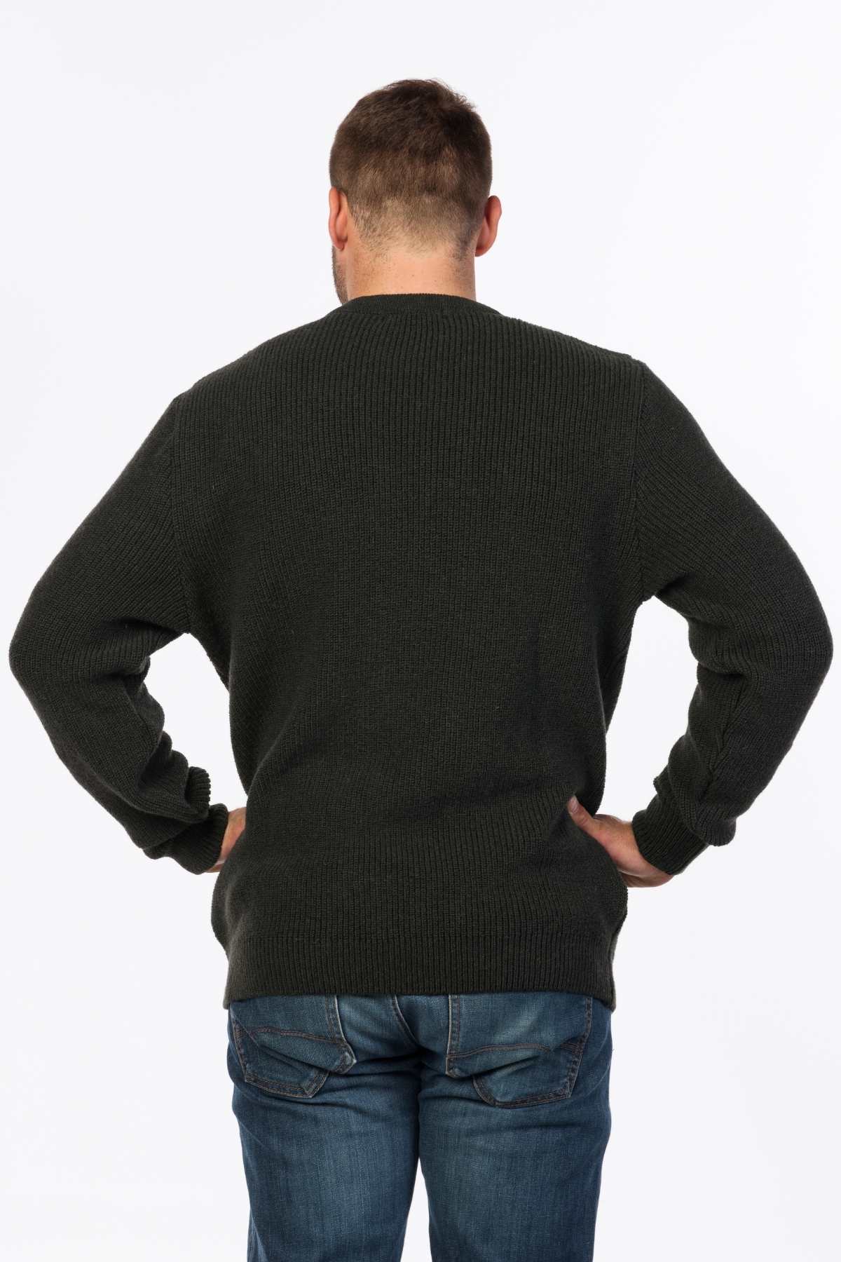 A person with short hair faces away, hands on hips, wearing an MKM Original Backyard Knit in dark green wool from New Zealand and blue jeans against a white background.