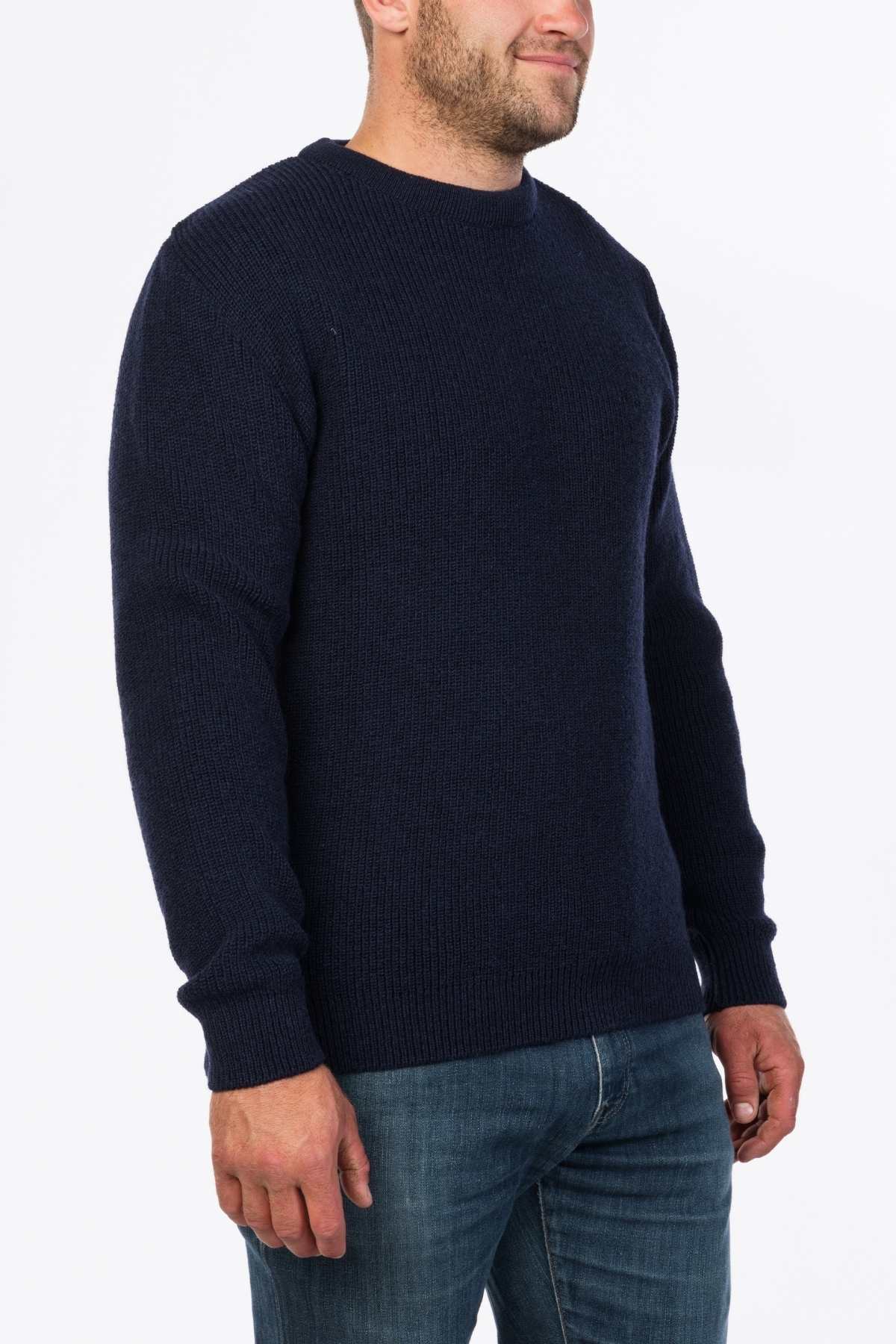 A person wearing the MKM Original Backyard Knit, a dark blue long-sleeve wool pullover made in New Zealand, and casual jeans stands against a white background. Their face is partially visible.