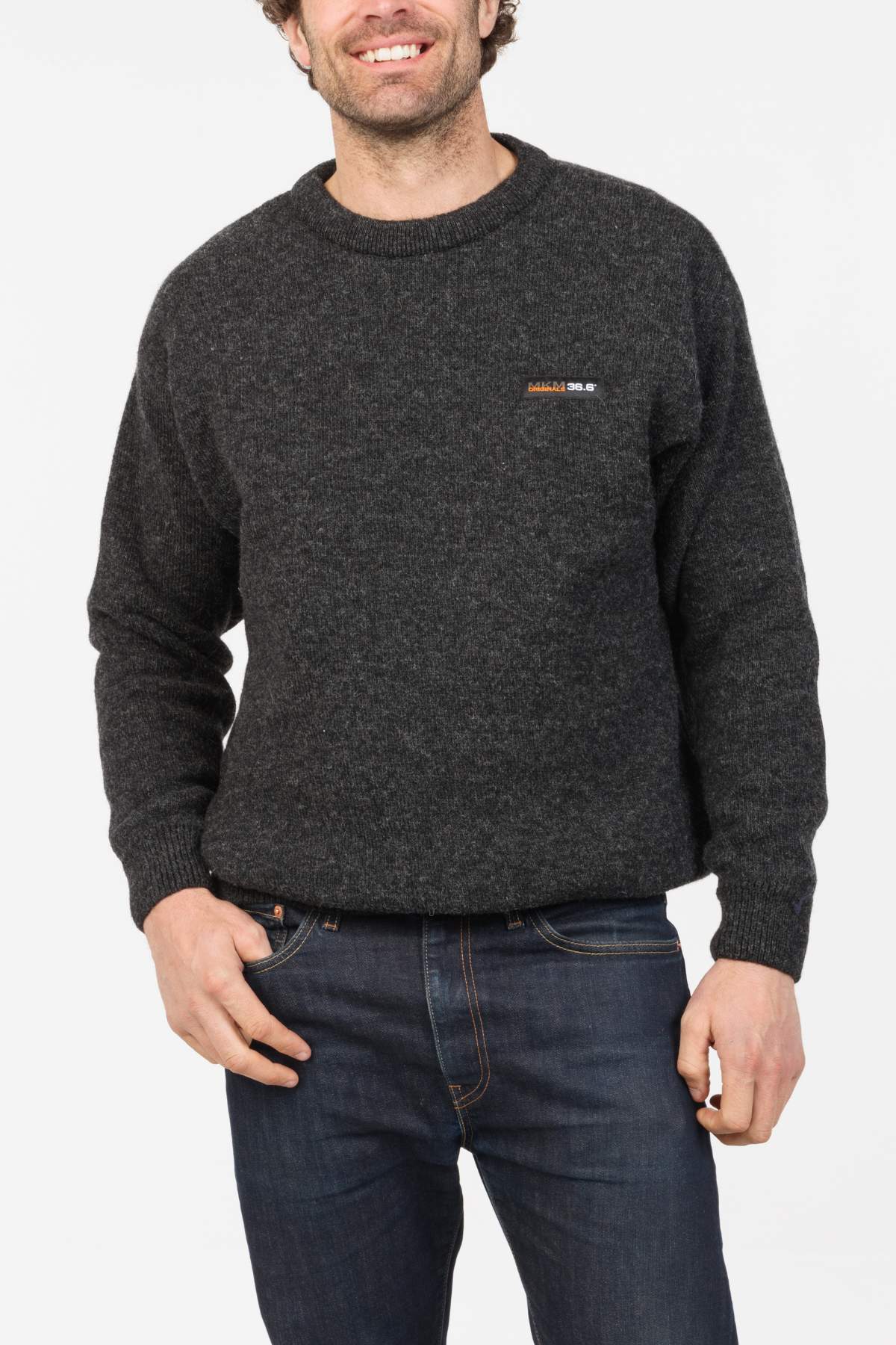 A man wearing the MKM Tasman Dual Layer Possum & Merino Crew in dark gray and blue jeans smiles with one hand in his pocket against a plain white background, highlighting outdoor performance and comfort.