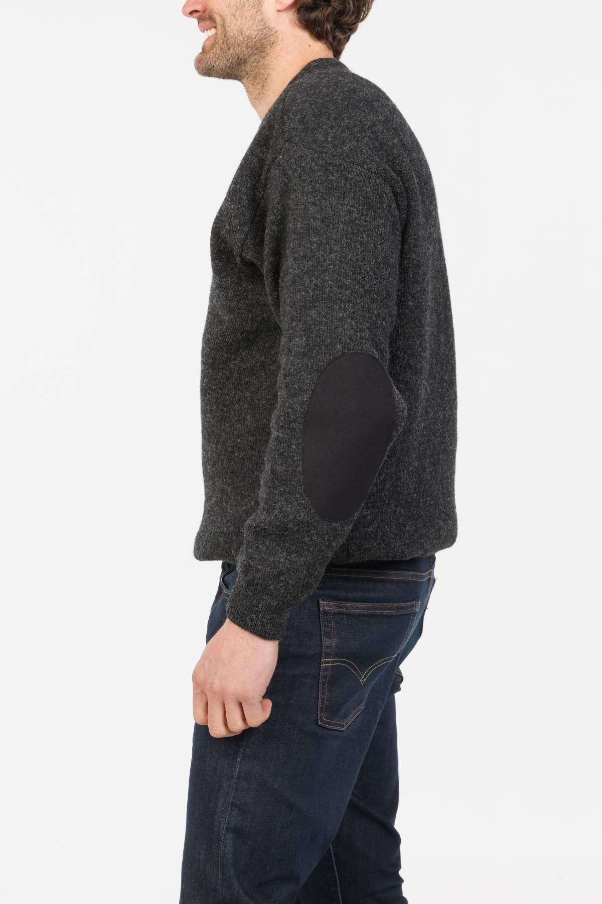 A man in profile wears the MKM Tasman Dual Layer Possum & Merino Crew sweater in dark gray with black elbow patches and dark blue jeans, standing against a plain white background.